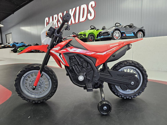 Vista laterale della moto elettrica Honda CRF 12V rossa con dettagli bianchi e logo Honda, in ambiente con sfondo di auto giocattolo colorate