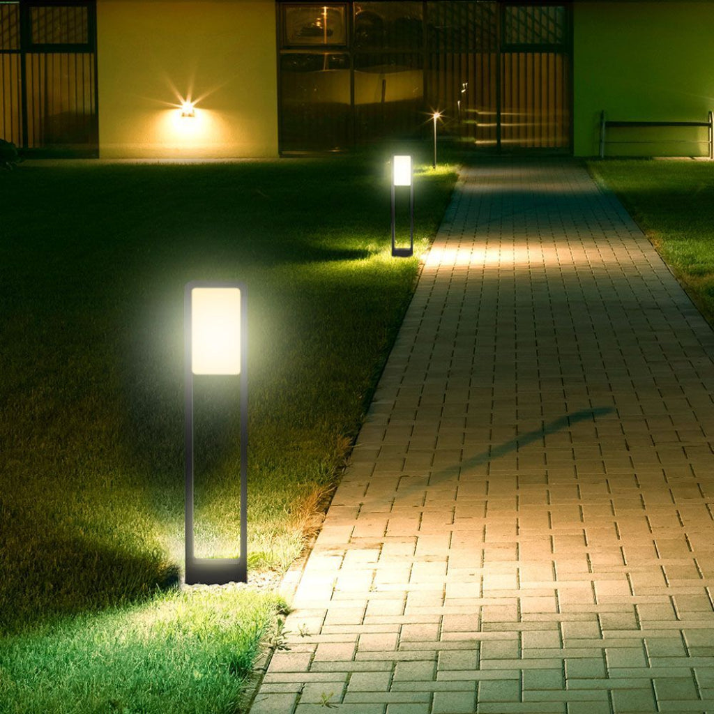 Immagine di lampade da esterno illuminate lungo un vialetto in giardino, con luce bianca calda e design minimalista in metallo nero.