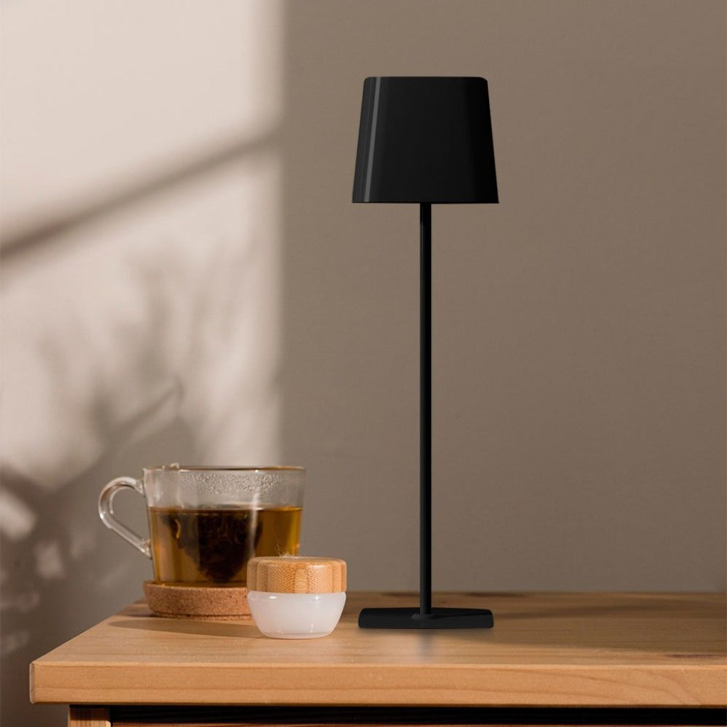 Lampada da tavolo nera moderna con base sottile e paralume a forma di cono, posizionata su un tavolo in un ambiente domestico con luce soffusa