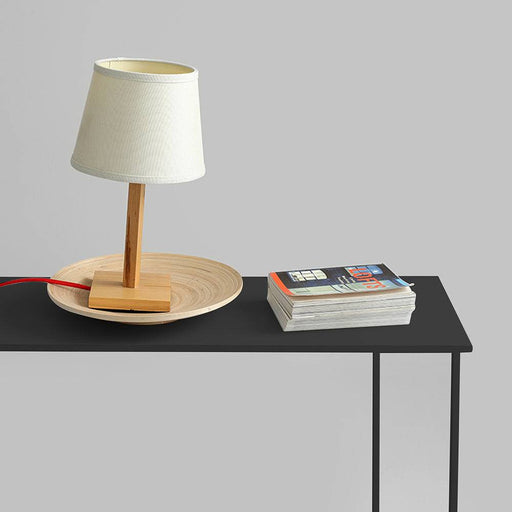 Lampada da tavolo in legno chiaro con paralume bianco, posizionata su una console nera con riviste, stile minimalista e funzionale