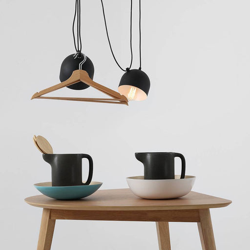 Luci sospese nere con cavi sottili, illuminate, sopra tavolo in legno con tazze e ciotole nere e bianche, stile scandinavo