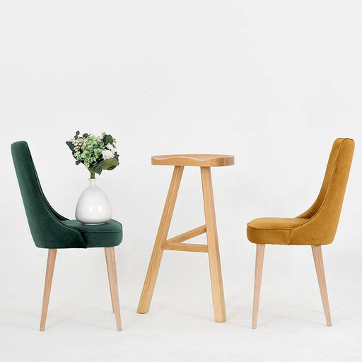 Sedia con rivestimento in velluto verde bottiglia, accompagnata da un tavolino in legno chiaro e una pianta con fiori bianchi, stile contemporaneo