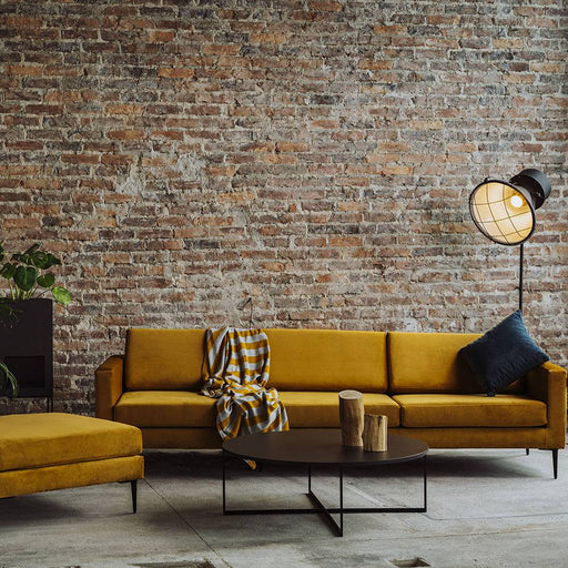 Divano in velluto color senape con schienale alto, stile moderno, posizionato davanti a parete in mattoni, atmosfera urbana.