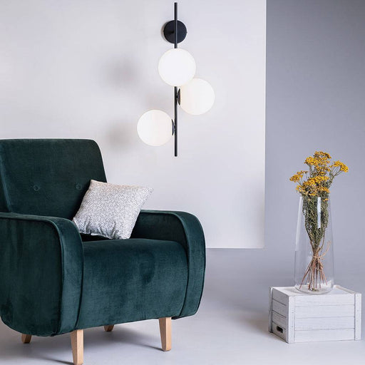 Lampada a parete BOBLER con tre globi di vetro bianco opaco e struttura nera, installata in soggiorno con parete bianca e divano verde scuro