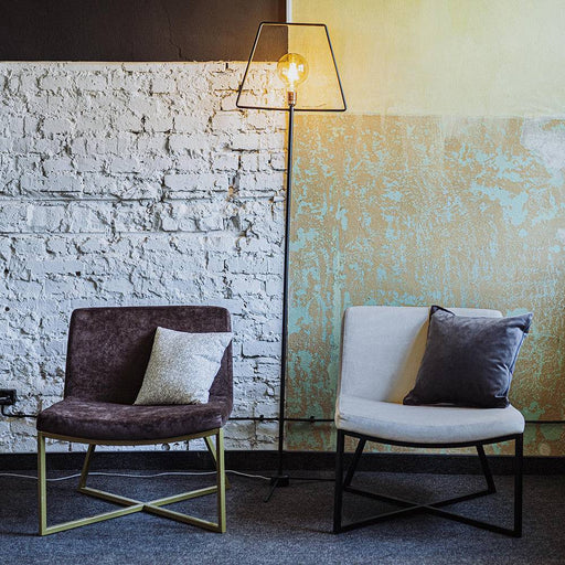 Angolo soggiorno con poltrona marrone in tessuto e base dorata, decorata con cuscino, in ambiente con pareti in mattoni bianchi
