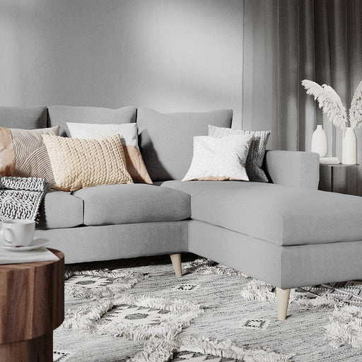Divano angolare in tessuto grigio con cuscini decorativi color beige e grigio, ambientato in soggiorno elegante con tappeto e decorazioni.