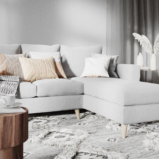 Divano angolare in tessuto grigio chiaro con cuscini decorativi beige e bianco, ambientazione elegante con pareti grigie e tappeto morbido, stile minimal.