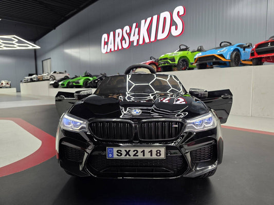 Retro dell'auto elettrica per bambini BMW M5 24 V nero metallizzato, con targa SX2118, dettagli lucidi, in un ambiente con altre auto giocattolo colorate.