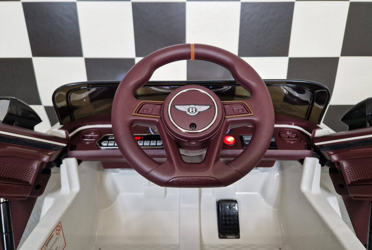 Interno dell'auto elettrica per bambini Bentley Bacalar, volante e pannello di controllo con luci e pulsanti, in ambiente con pavimento a scacchi.