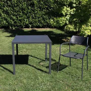 Tavolo Manchester impilabile 80 X 80 grigio scuro in acciaio verniciato a polvere, posizionato in un giardino con sedia abbinata.