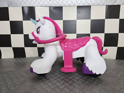 Vista frontale del cavallo a dondolo unicorno bianco con dettagli rosa e corno blu, sedile rosa e ruote nere, su sfondo a scacchi