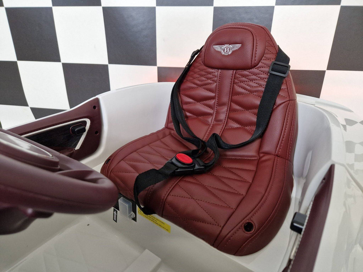 Sedile in pelle rossa con cuciture trapuntate e logo Bentley, parte di un'auto elettrica per bambini, con cinturino di sicurezza nero e maniglia laterale, sfondo a scacchi bianco e nero.