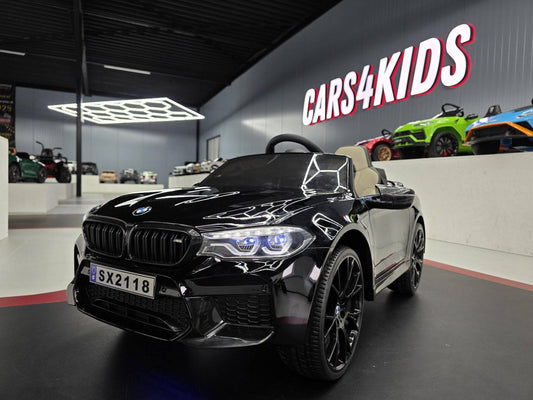 Auto elettrica per bambini BMW M5 24 V nero metallizzato, dettagli realistici, sedile in beige, in un showroom con auto giocattolo colorate sullo sfondo.