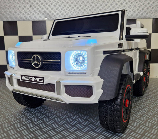 Auto elettrica per bambini Mercedes G63 AMG 6x6 bianca con fari LED accesi, dettagli realistici e grandi ruote nere con bordi rossi.