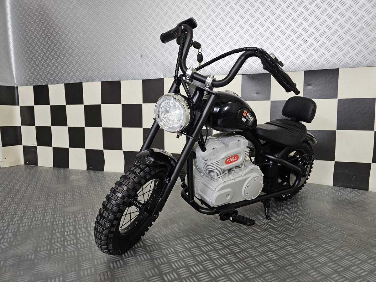 Vista laterale della moto elettrica per bambini modello chopper nero con dettagli in stile motociclista, grande ruota anteriore e motore visibile.