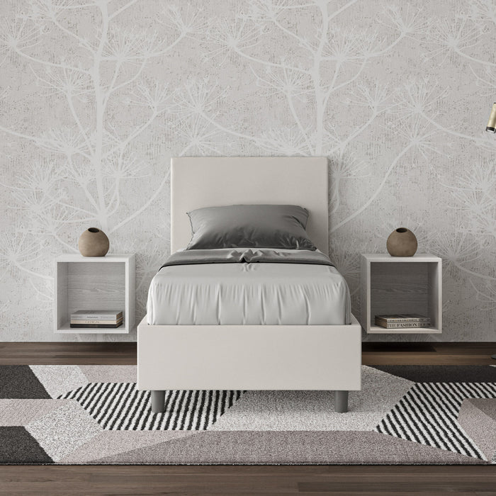 Letto singolo Adele bianco in similpelle con testiera imbottita, inserito in una camera moderna con comodini sospesi e tappeto geometrico.