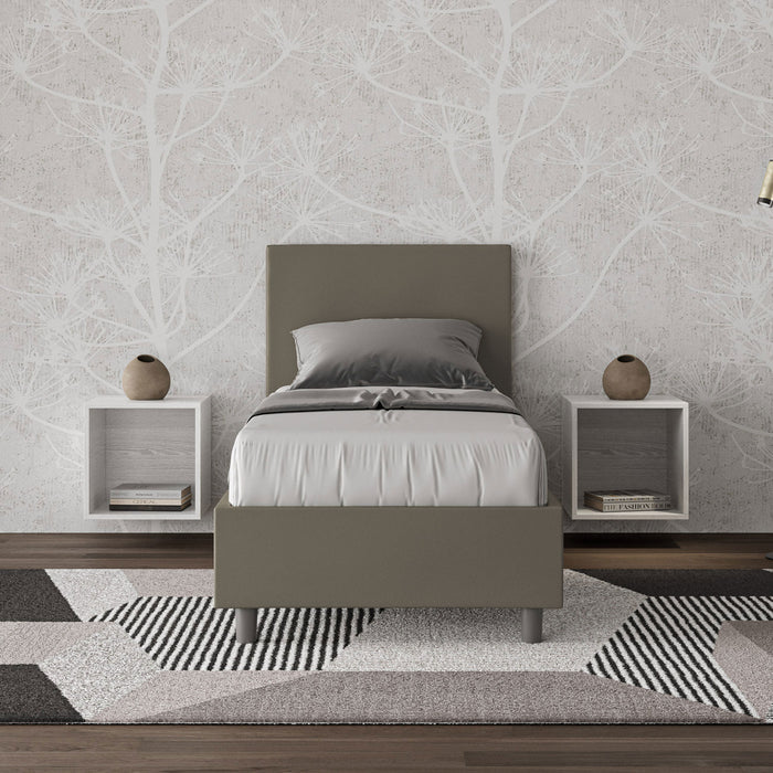 Letto singolo Adele grigio-verde in similpelle con testiera imbottita, ambientato in camera moderna con tappeto geometrico e comodini sospesi.