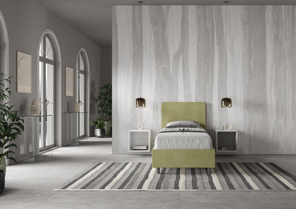 Letto singolo imbottito Adele 80x210 verde salvia in microfibra, inserito in ambiente elegante con tappeto a righe e grandi finestre.