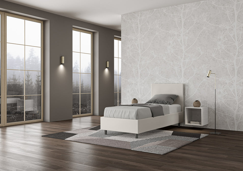 Letto singolo Adele bianco in similpelle con struttura imbottita, posizionato in una stanza moderna con pavimento in legno e grandi finestre.