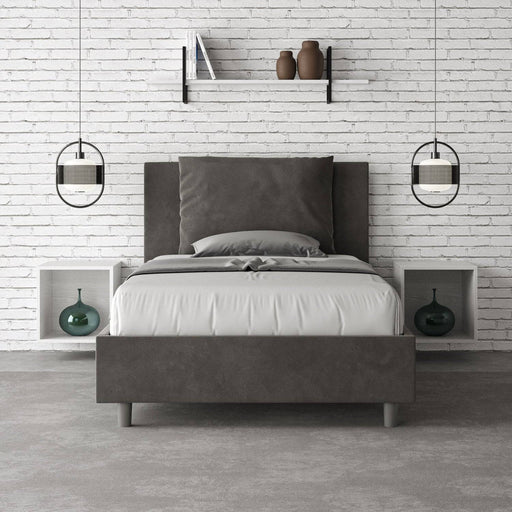 Letto singolo Antea 100x200 in microfibra grigia con contenitore, inserito in una camera moderna con parete in mattoni bianchi e arredi minimal.