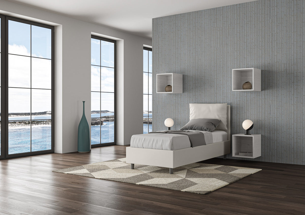 Letto singolo Antea bianco in similpelle con contenitore, posizionato in una camera con ampie finestre e pavimento in legno.