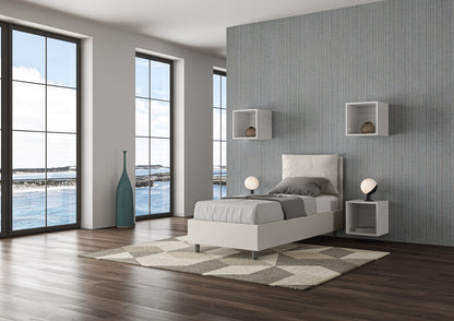 Letto singolo Antea bianco in similpelle con contenitore, posizionato in una camera con ampie finestre e pavimento in legno.