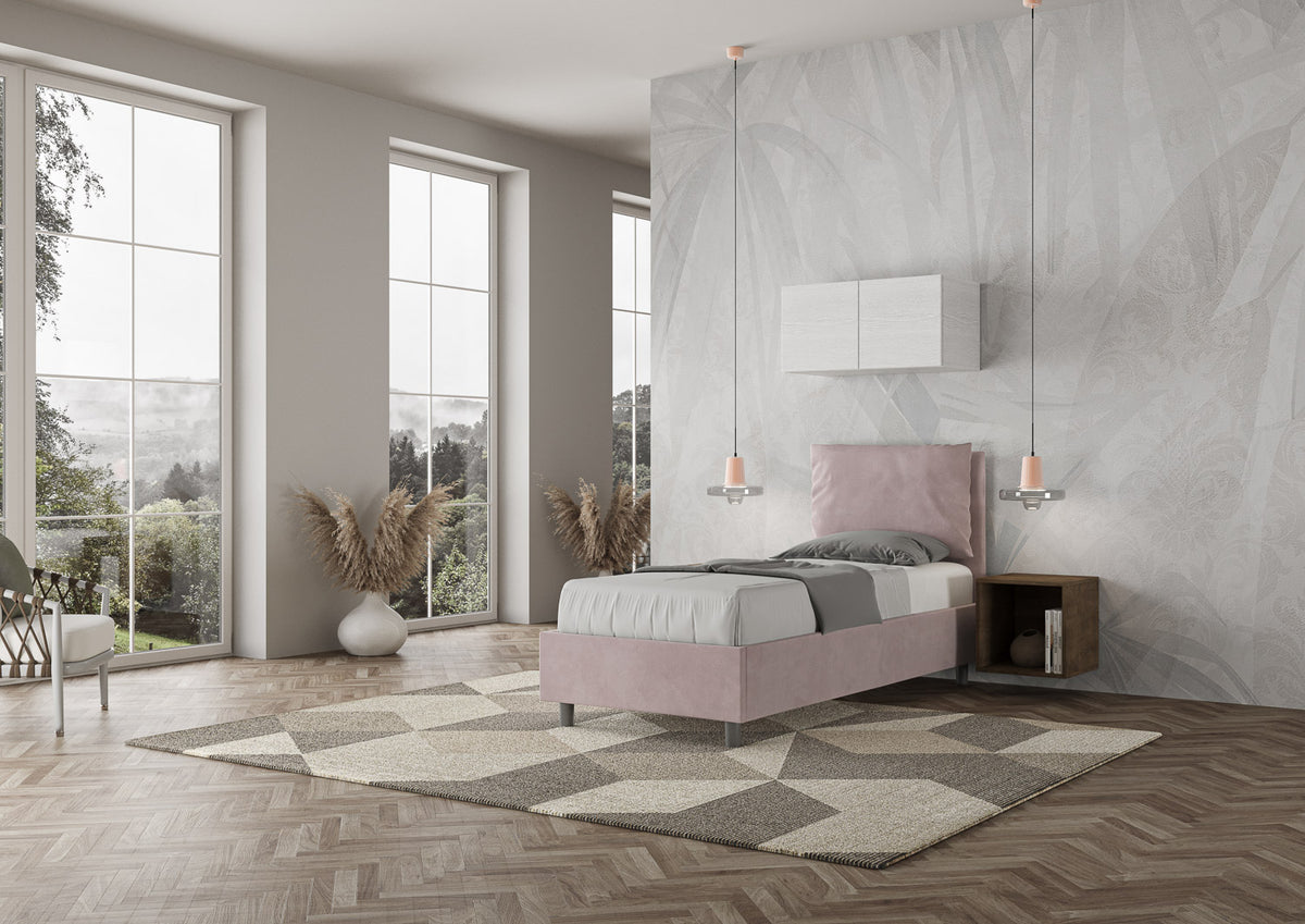 Letto Antea rosa 80x190 con struttura imbottita in microfibra, ambientato in una stanza moderna con parquet e grandi finestre.