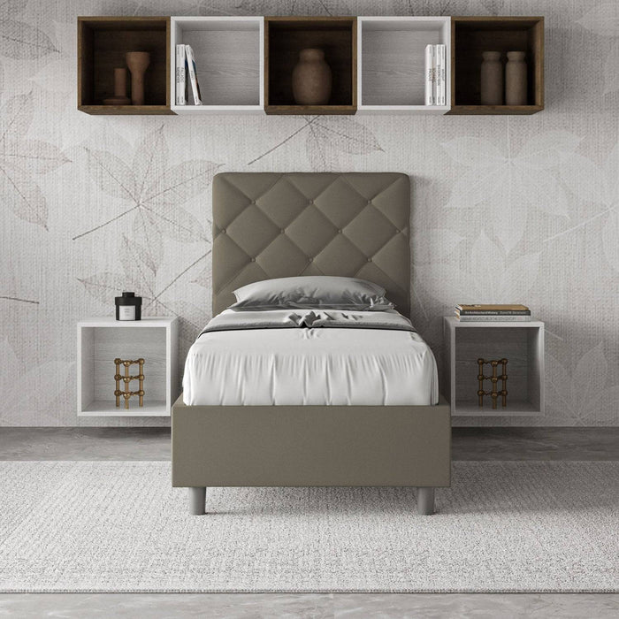 Letto singolo Priya grigio in similpelle con testiera trapuntata, inserito in una camera moderna con mensole e comodini sospesi.