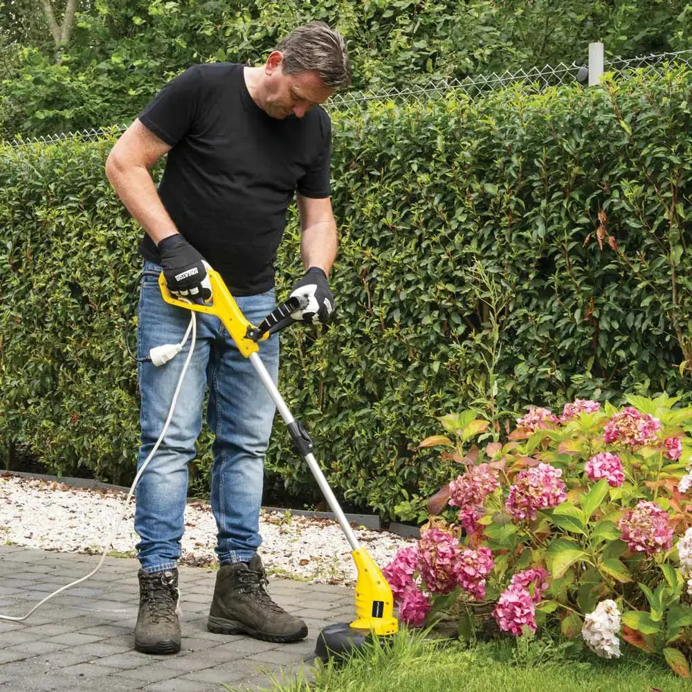 Uomo che utilizza il tagliabordi elettrico Powerplus 350W con guanti, in giardino con cespugli e fiori, per potatura e rifinitura del prato