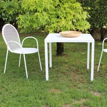 Tavolo Manchester impilabile 80 X 80 bianco in acciaio verniciato a polvere, ambientato in un giardino con sedie coordinate.