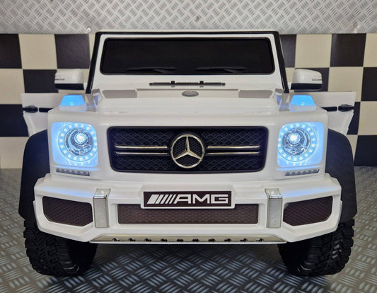 Vista frontale dell'auto elettrica per bambini Mercedes G63 AMG 6x6 bianca con dettagli come il logo Mercedes e fari LED luminosi.