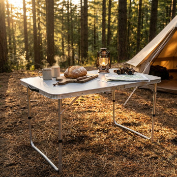 Tavolo pieghevole in MDF e alluminio bianco, utilizzato all'aperto tra alberi, con cibo, tazze e lanterna per attività outdoor