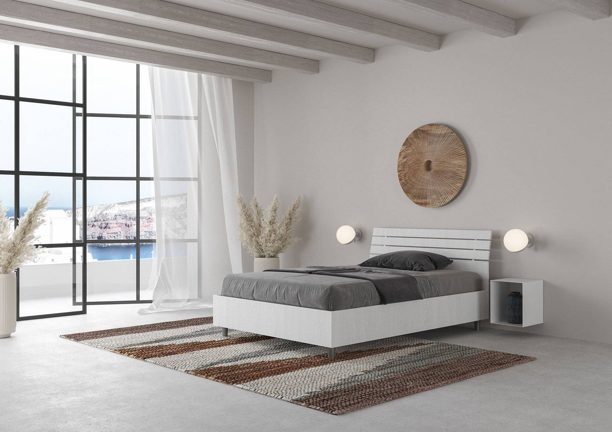 Letto singolo Ankel bianco con contenitore, posizionato in una camera con ampie finestre e decorazioni naturali.